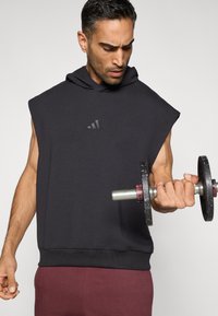 Tamsiai pilkas bliuzonas be rankovių su priekiu esančiu logotipu, striukės apačia su dantytu kraštu ir raumeningas asmuo, laikantis juodą hantelį treniruočių pozoje.