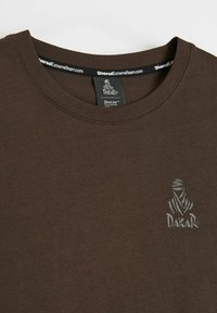 Ciemnobrązowa koszulka z dekoltem w serek, z małym szarym logo Dakar na lewej piersi oraz czarną wewnętrzną metką na karku z napisem DiverseExtremeTeam.com.