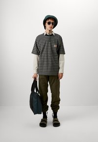 Polo rayé gris et bleu marine, superposé à une chemise blanche à manches longues. Pantalons cargo vert olive, sac gris, sandales foncées, chapeau de seau vert.