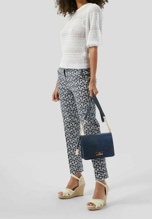 Hvid strikket top med korte ærmer, mønstrede marineblå og hvide bukser, beige kilehæle, og en marineblå tekstureret håndtaske med guldaccentuer.