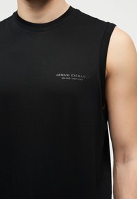 Chemise noire sans manches portée par une personne, arborant un petit texte "Armani Exchange Milano / New York" sur la poitrine.