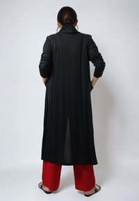 Lange zwarte cardigan met een hoge kraag en geribbeld textuur, gedragen over rode wijde broek. Heeft een split achter voor beweging.