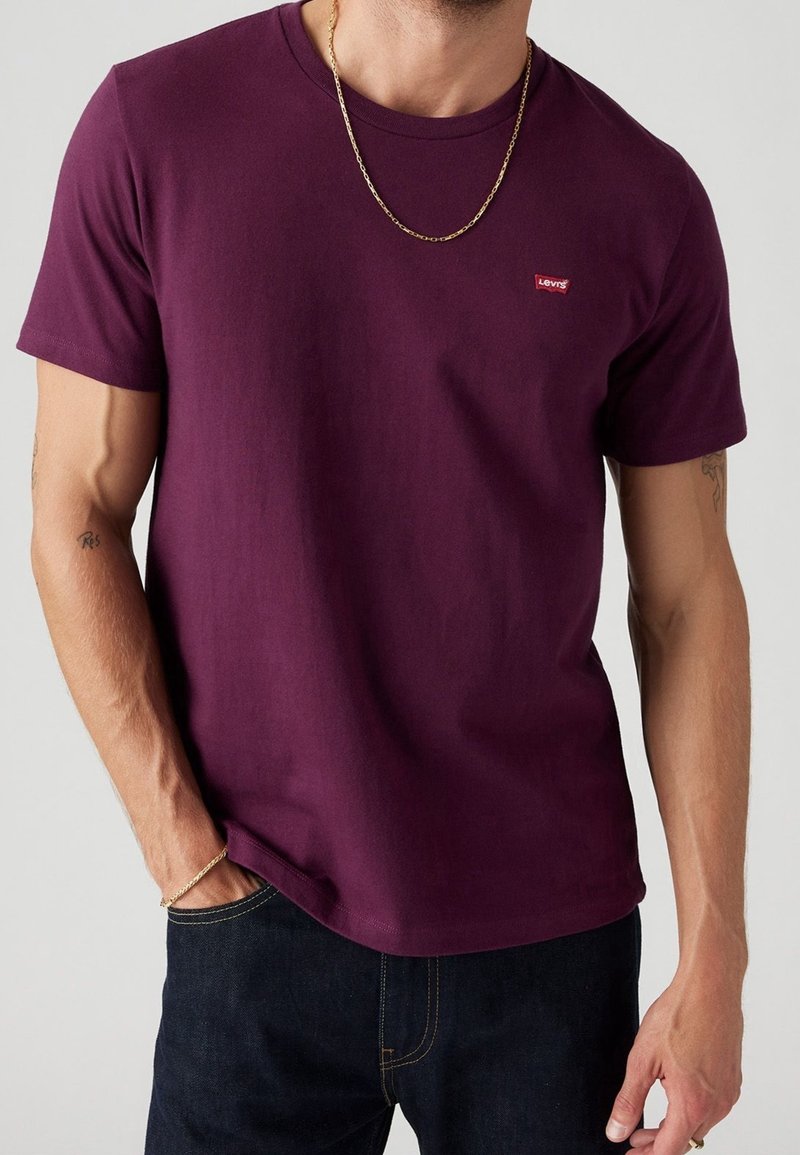 T-shirt manches courtes bordeaux en coton, avec un petit logo rouge Levi's sur la poitrine, associé à un jean foncé et une chaîne en or.