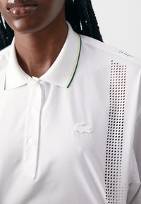 Bílá polo košile s límečkem na knoflíky, vertikálním perforovaným detailem na jedné straně a malým bílým logem Lacoste na hrudi.