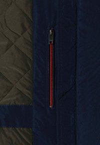 Vatteret stof med diamantsting i mørk oliven og marineblå, med en lodret marineblå lynlås med røde tænder og en sølv trækstang.