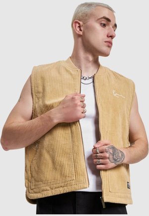 Gilet en velours côtelé beige avec une fermeture éclair à l'avant, des poches latérales et un petit logo. Il présente un col lisse et une texture côtelée.