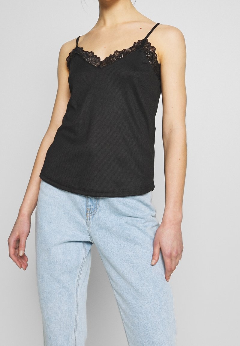 Camisola nera con bordo in pizzo lungo il colletto. Spalline sottili regolabili, texture a costine morbida, design aderente, indossata con jeans azzurri chiari.