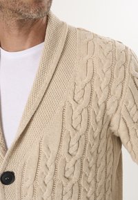 Beige strikket cardigan med kabelmønster, struktureret væv, stor sjalkrave og en sort knap. Båret over en hvid t-shirt.