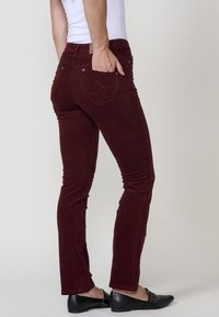 Persoon in bordeaux corduroy broek en zwarte instappers, met hand in de achterzak, tegen een effen lichte achtergrond.