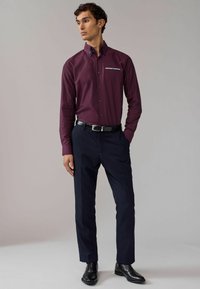 Camisa burdeos de manga larga con cuello puntiagudo, que presenta un parche decorativo y detalles en azul marino; combinada con pantalones navy a medida y zapatos negros.