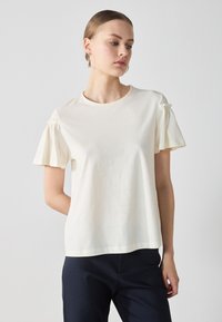 T-shirt a maniche corte in cotone crema con dettagli arricciati sulle maniche, scollatura rotonda e vestibilità rilassata. Texture liscia e design minimal.