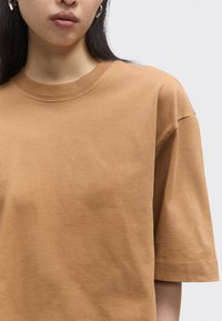 Woman wearing plain brown short-sleeve crew neck T-shirt with gold hoop earrings, shown from shoulders to lips against a white background.