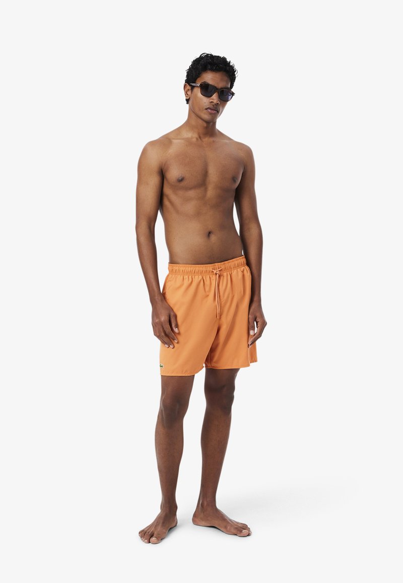 Jonge man draagt oranje zwembroek en zwarte zonnebril, staat blootsvoets tegen een effen witte achtergrond.