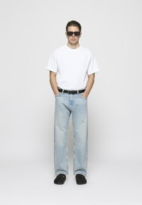 Camiseta blanca de manga corta, jeans de color azul claro, cinturón negro y zapatos negros. Modelo con gafas de sol rectangulares oscuras, de pie.