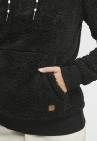 Sudadera con capucha de felpa negra con una superficie texturizada, capucha con cordón, bolsillo canguro y un dobladillo negro acanalado. Presenta una pequeña etiqueta de marca.