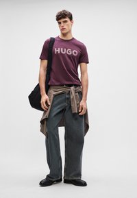 Maroon t-särk, millel on heledat halli värvi "HUGO", koos lõdva, pleekinud siniste teksade ja seotud beeži särgiga, kandes musta õlakotti.