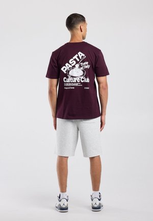 Man draagt een donkerpaars T-shirt met witte pastagrafiek en tekst, lichtgrijze korte broek, witte sokken en witte sneakers, staand met de rug naar voren.
