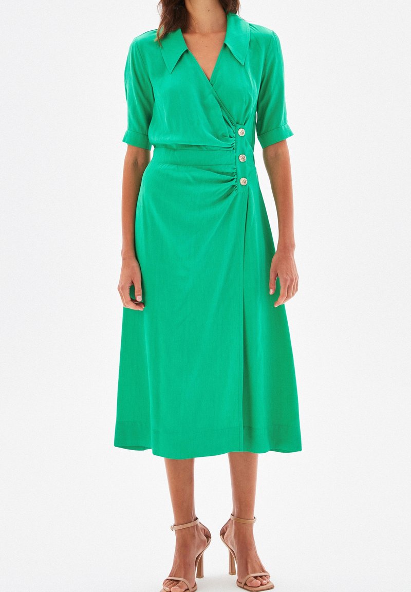 Robe portefeuille verte jusqu'aux genoux avec manches courtes, un col, et des boutons décoratifs. Tissu lisse avec un devant drapé et une silhouette simple.