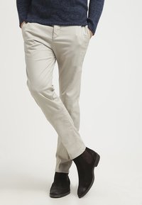 Persona che indossa chinos beige e stivali Chelsea in suede nero, in piedi con le gambe incrociate e le mani in tasca.