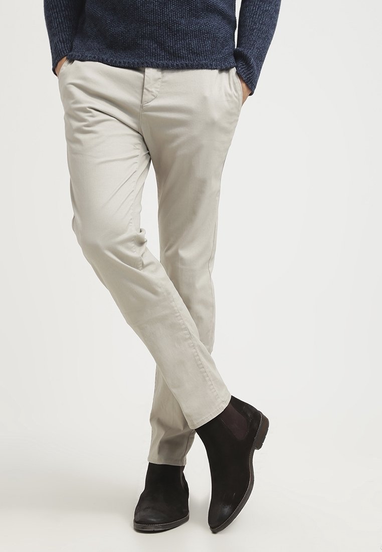 Persona che indossa chinos beige e stivali Chelsea in suede nero, in piedi con le gambe incrociate e le mani in tasca.