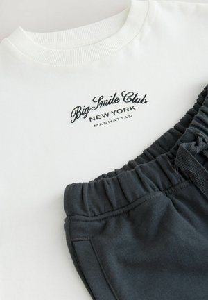Balta apģērba kreklā ar melnu izšūtu tekstu "Lielā Smaida Klubs Ņujorkā Manhetenā" blakus tumši pelēkiem sporta biksēm ar elastīgu jostasvietu un kabatām.