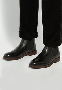 Black leather Chelsea boots with elastic side panels and a stacked wooden heel. Worn with rolled black corduroy pants, showcasing ankle detail.