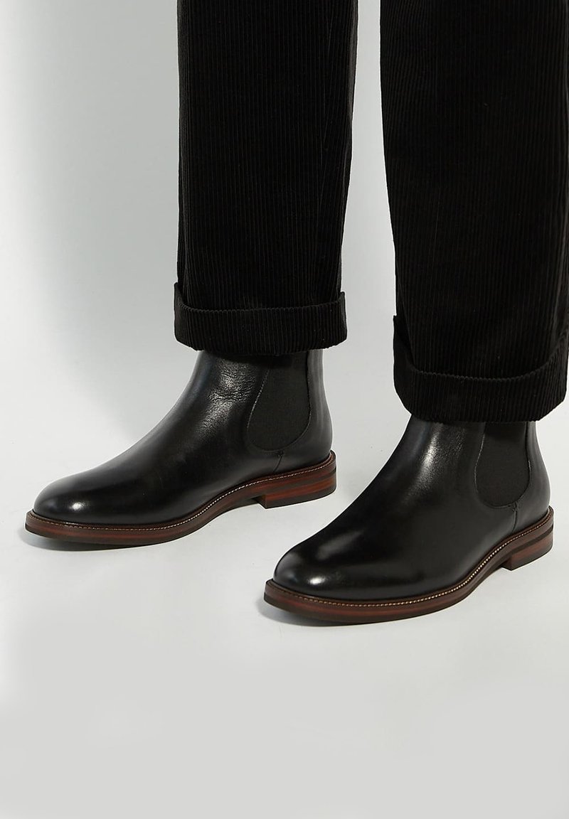 Black leather Chelsea boots with elastic side panels and a stacked wooden heel. Worn with rolled black corduroy pants, showcasing ankle detail.