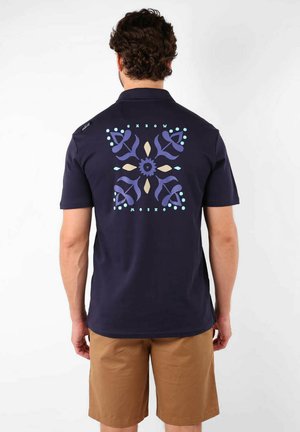 Chemise à manches courtes bleu marine avec un graphique géométrique coloré au dos. Fabriquée en tissu doux avec un col classique et des accents de boutons.