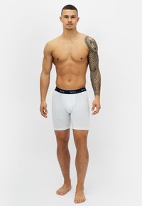 Valkoiset puuvillabokserit, joissa on tumma vyötärö, ja niissä on logo. Mallilla on lyhyet hiukset ja täysikokoinen käsivarteen tehty tatuointi.