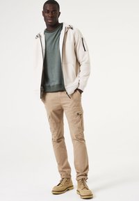 Sweat à capuche zippé beige clair avec des accents noirs, sweatshirt vert à col rond, pantalon cargo beige, et bottes lacées marron clair.