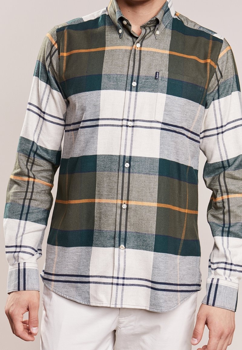 Homme portant une chemise à carreaux à manches longues boutonnée avec des rayures vertes, blanches, grises et orange, associée à un pantalon blanc.