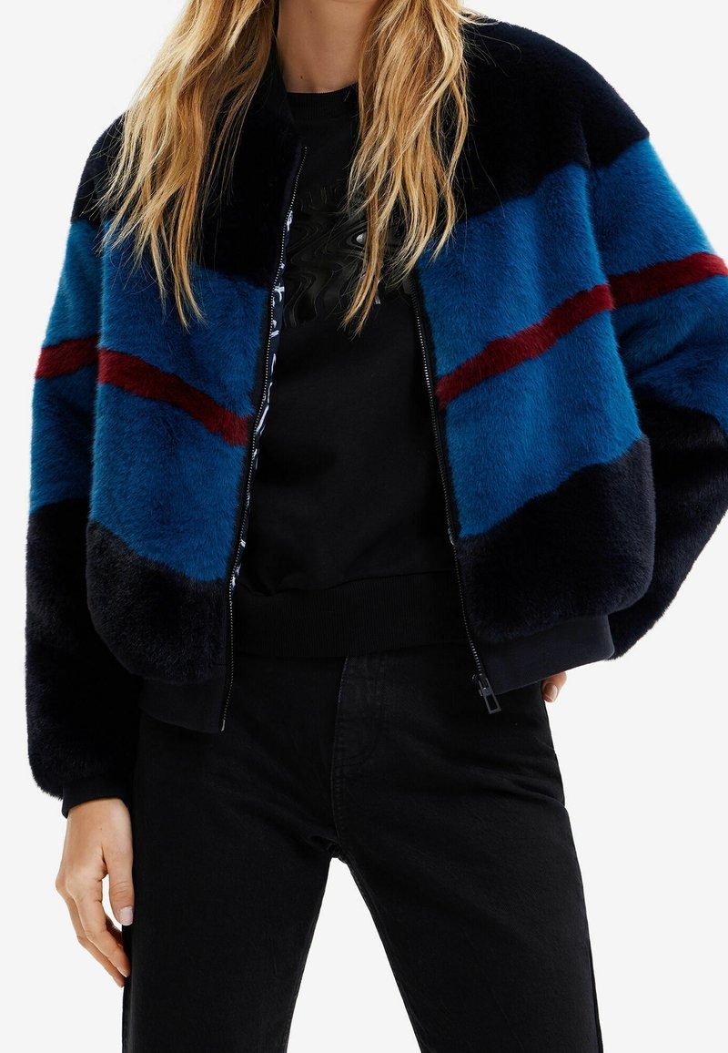 Veste bomber en fausse fourrure avec des rayures bleues, noires et bordeaux, devant zippé et poignets côtelés, portée sur un sweat-shirt noir.