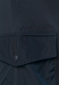 Svart textilprodukt med en slät stickad yta som har en kontrasterande glansig ficka, präglat varumärke och tryckknappdetalj.