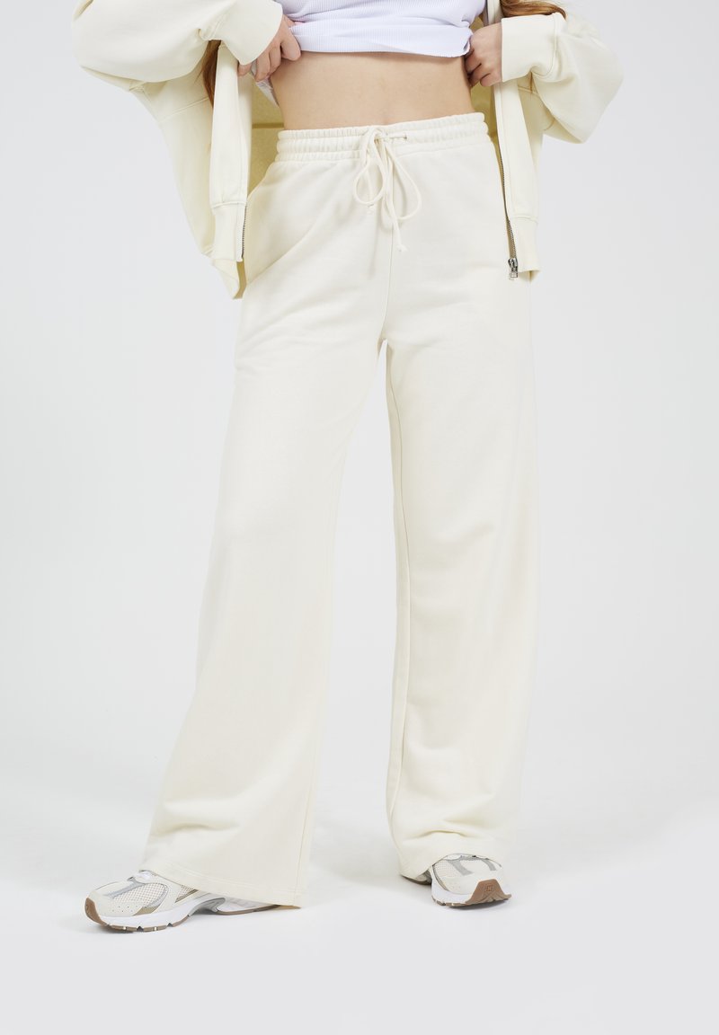 Crèmewitte, wijde sweatpants met een elastische tailleband en trekkoord. Zachte textuur, gladde afwerking, gecombineerd met witte en grijze sneakers.