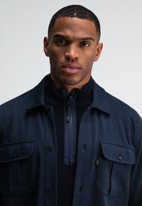 Marineblaues Button-up-Jacket mit zwei Brusttaschen, über einem zipfeligen Pullover getragen. Glattes Material mit strukturiertem Kragen und schulterlangen Ärmeln.