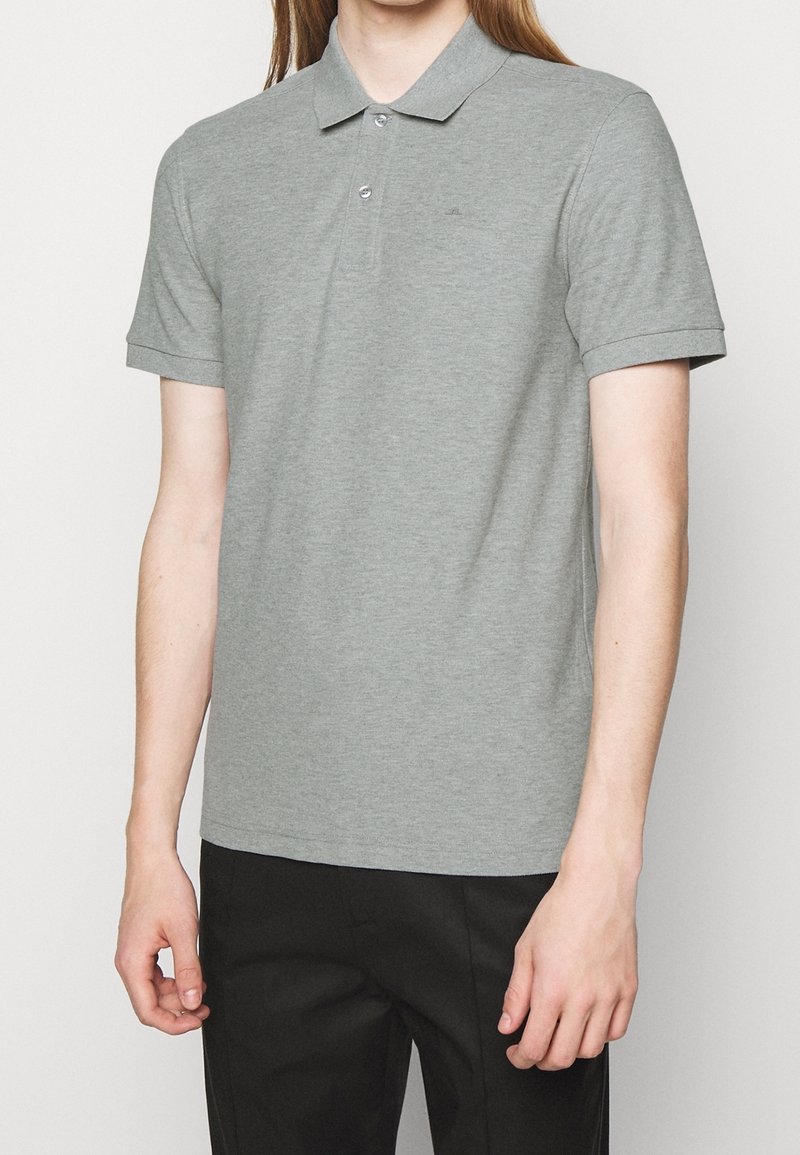 Personne portant un polo gris à manches courtes avec deux boutons et un pantalon noir, debout devant un fond blanc uni.
