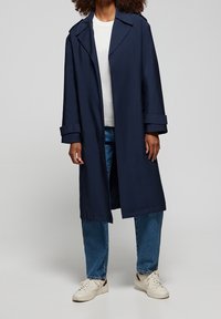 Osoba ubrana w granatowy długi płaszcz na białej koszuli, niebieskich dżinsach i białych trampkach, stojąca na tle jednolitego szarego tła.