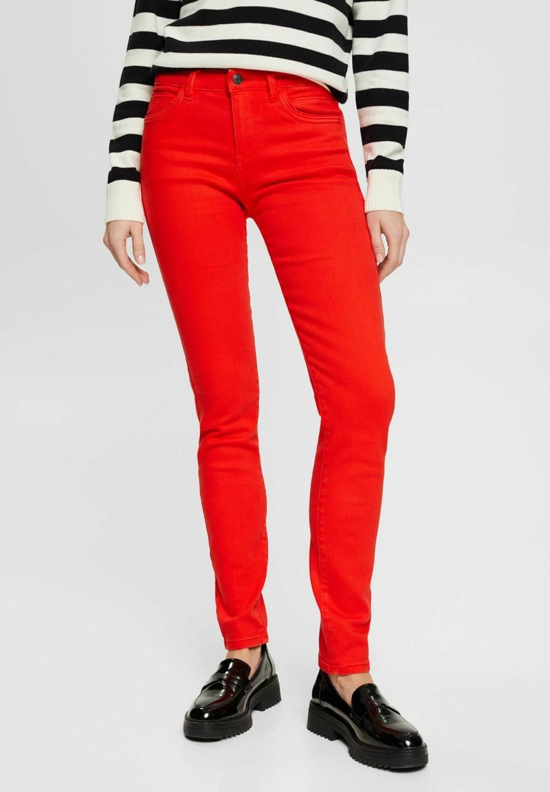 Personne portant un jean slim rouge vif, des mocassins noirs brillants et un haut rayé noir et blanc à manches longues, debout sur un fond uni.