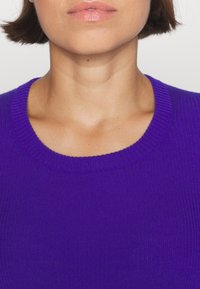Fioletowy sweter dzianinowy o prążkowanej teksturze i okrągłym dekolcie, z gładkiego materiału o delikatnym połysku. Zbliżenie na odzież.