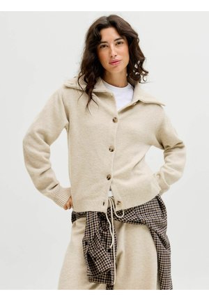 Gilet beige realizzato in materiale morbido, caratterizzato da un colletto, bottoni frontali e polsini a coste. Indossato sopra pantaloni a quadri.