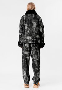 Persona vestida con una chaqueta y pantalones texturizados en negro y gris, con collar y puños de pelo negro, de pie de espaldas, zapatos de plataforma en beige visibles.