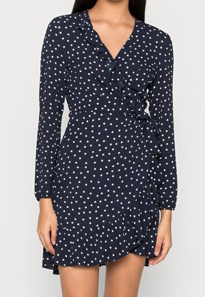 Femme portant une robe portefeuille bleu marine à pois blancs avec des bords froncés, manches longues et un décolleté en V.