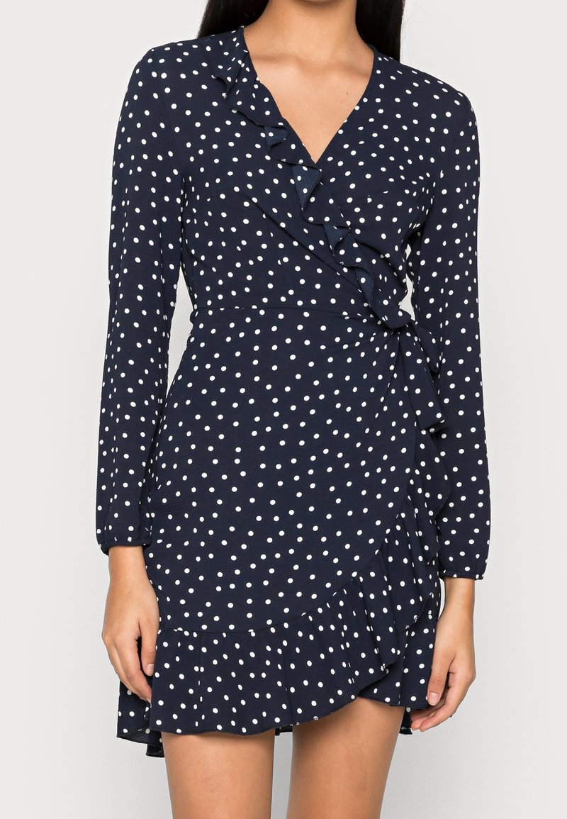 Femme portant une robe portefeuille bleu marine à pois blancs avec des bords froncés, manches longues et un décolleté en V.