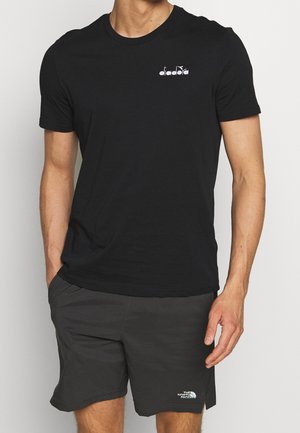 Homme portant un T-shirt Diadora noir uni et un short gris foncé The North Face, debout avec une main dans la poche.