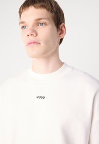Valge dressipluus ümmarguse kaeluse ja musta "HUGO" logoga rinnal. Sile tekstuur ja rahulik sobivus.