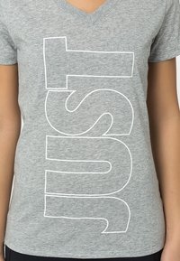 Camiseta de algodón gris con cuello en V; presenta un texto en blanco en negrita que dice "JUST" en orientación vertical. Textura suave, diseño ajustado.