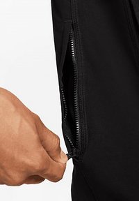 Black athletic pants featuring a zippered pocket. Close-up view shows a hand gripping the zipper pull, revealing smooth fabric and stitching details.