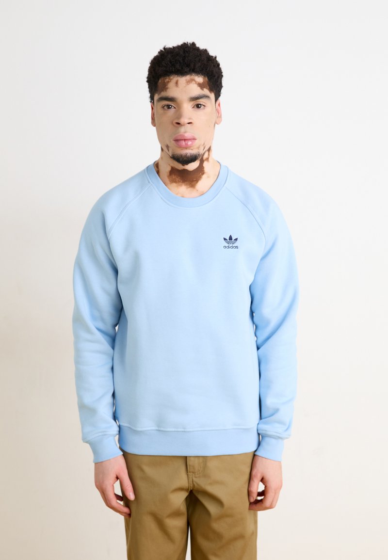 Svetlo modra majica s kapuco, z rebrastimi manšetami in robom, na prsih ima majhen črn Adidas logo, v kombinaciji s bež hlačami.