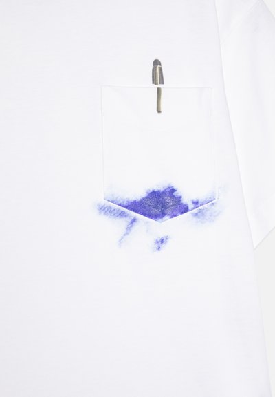 Camiseta blanca con un diseño de manchas de pintura azul cerca del fondo y un bolsillo en el pecho con un bolígrafo guardado dentro.