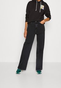 Felpa nera con cappuccio e tasca frontale, abbinata a jeans neri a vita alta. Le calzature presentano dettagli in teal e una suola leggera.